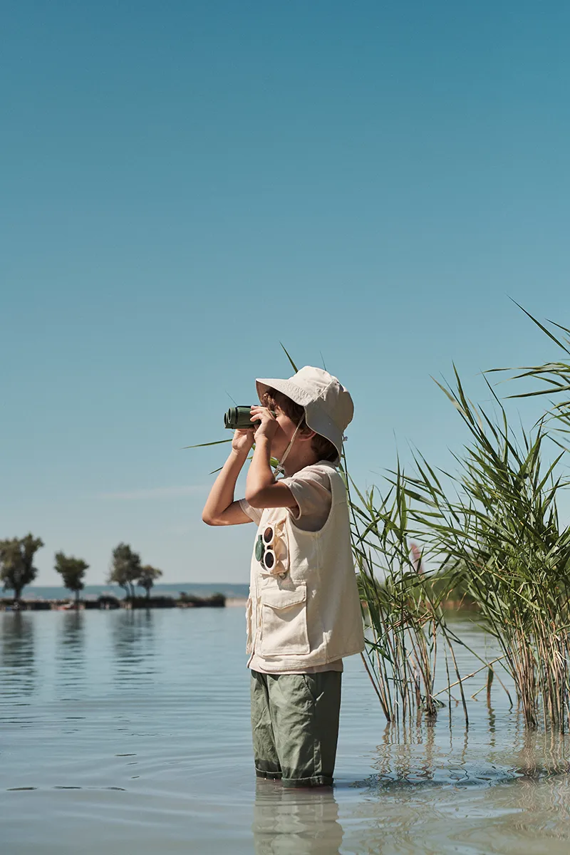 Ein Kind, das einen Sonnenhut und eine Weste trägt, steht im flachen Wasser in der Nähe von hohem Schilf und blickt durch ein Fernglas auf den blauen Himmel und die entfernten Bäume auf der anderen Seite des Sees.