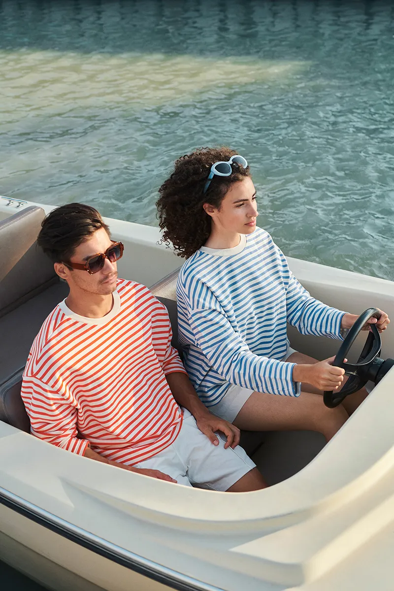 Zwei Personen sitzen in einem kleinen Boot auf dem Wasser. Einer trägt ein rot-weiß gestreiftes Hemd und eine Sonnenbrille, der andere ein blau-weiß gestreiftes Hemd und weiße Shorts und steuert das Boot. Beide sehen entspannt und konzentriert aus.